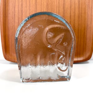 Puede incluir: Una escultura de vidrio transparente de un elefante con interior marrón. El elefante tiene una cara sonriente y está sobre un fondo de madera. La escultura mide aproximadamente 10 cm de altura.