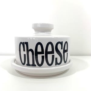 Puede incluir: Plato de queso de cerámica blanca con tapa. El plato tiene la palabra "Cheese" impresa en negro en la parte delantera.