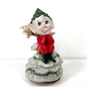 Puede incluir: Una figura de cerámica de un elfo con un sombrero puntiagudo verde, orejas grandes y un rostro pálido. El elfo viste un traje rojo brillante con botas negras y está sentado sobre una formación rocosa texturizada de color gris claro.