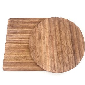 Può includere: Due vassoi da portata in legno, uno rotondo e uno rettangolare. Entrambi i vassoi hanno una venatura del legno e sono realizzati in legno marrone chiaro.