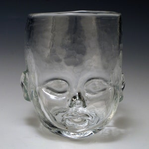 May include: A clear glass drinking glass shaped like a baby's face. The glass has a rounded bottom and a wide opening.