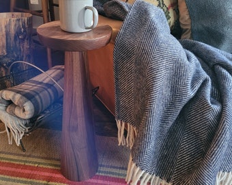 May include: A wooden side table with a dark brown finish and a rounded top. A white enamel mug sits on the table. A blue and white patterned throw blanket is draped over a chair in the background.