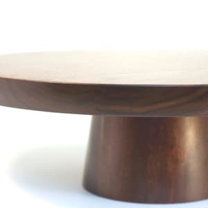 May include: A round wooden cake stand with a dark brown finish. The stand has a wide, flat top and a tall, cylindrical base.