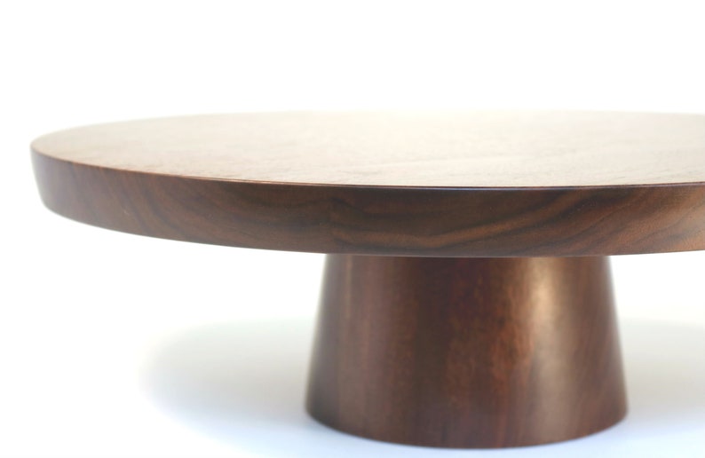 May include: A round wooden cake stand with a dark brown finish. The stand has a wide, flat top and a tall, cylindrical base.
