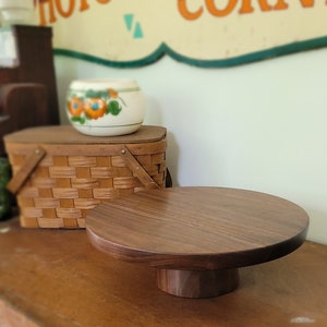 May include: A round wooden cake stand with a dark brown finish. The stand has a wide base and a smaller top, perfect for displaying cakes, pastries, or other treats.