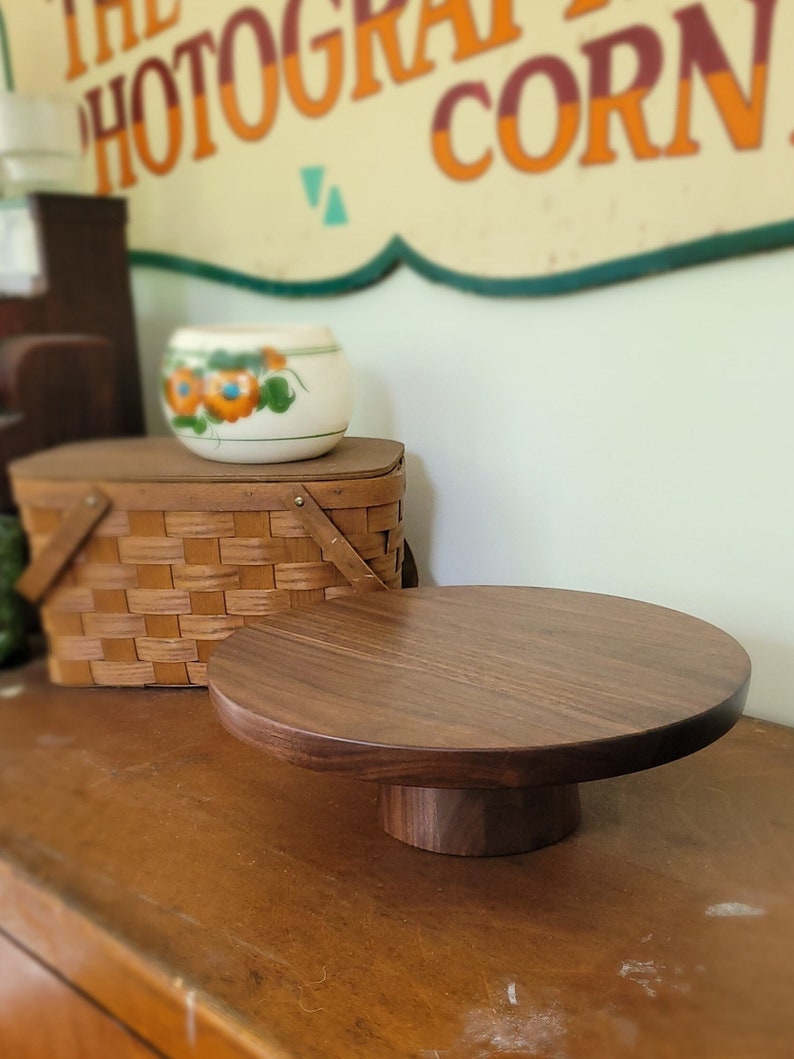 May include: A round wooden cake stand with a dark brown finish. The stand has a wide base and a smaller top, perfect for displaying cakes, pastries, or other treats.