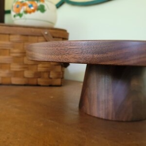 May include: A round wooden cake stand with a dark brown finish. The stand has a wide base and a narrow top.