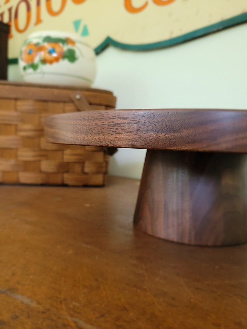 May include: A round wooden cake stand with a dark brown finish. The stand has a wide base and a narrow top.