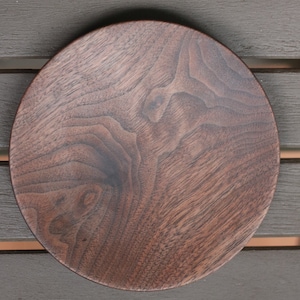 May include: A round, dark brown wooden plate with visible wood grain. The plate is made of a single piece of wood and has a smooth, polished surface. The plate is sitting on a dark brown slatted surface.
