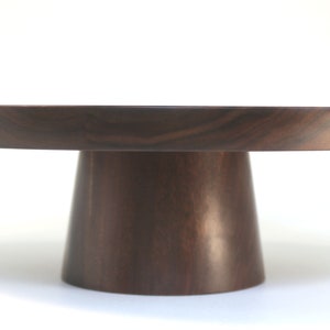 May include: A round wooden cake stand with a dark brown finish. The stand has a wide base and a narrow top, making it perfect for displaying cakes and other desserts.