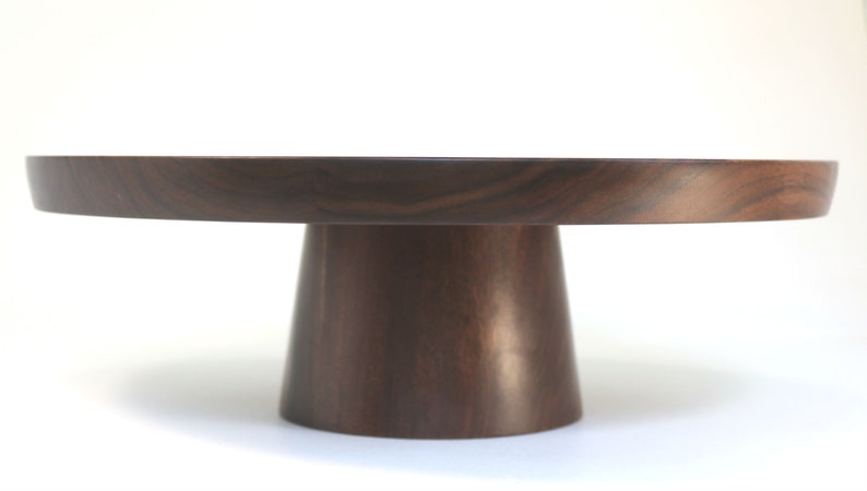 May include: A round wooden cake stand with a dark brown finish. The stand has a wide base and a narrow top, making it perfect for displaying cakes and other desserts.