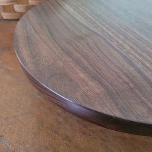 May include: A dark brown wooden cake stand with a smooth, rounded top and a slightly raised edge. The stand is made of solid wood and has a natural wood grain.