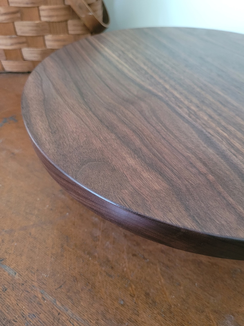 May include: A dark brown wooden cake stand with a smooth, rounded top and a slightly raised edge. The stand is made of solid wood and has a natural wood grain.