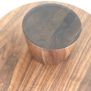 May include: A round wooden serving tray with a smaller round wooden pedestal in the center. The tray and pedestal are both made of dark wood with a natural grain pattern.