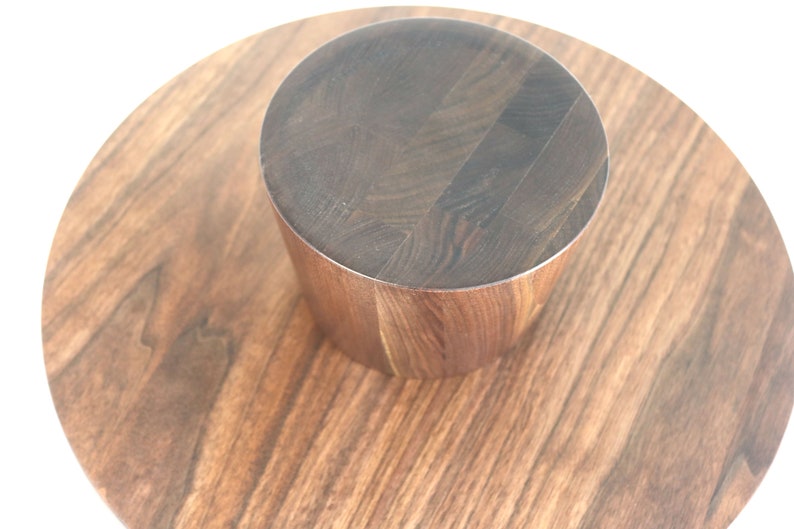 May include: A round wooden serving tray with a smaller round wooden pedestal in the center. The tray and pedestal are both made of dark wood with a natural grain pattern.