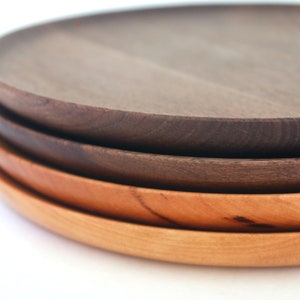 May include: A stack of four wooden plates in varying shades of brown. The top plate is a dark, rich brown, followed by lighter shades of brown and reddish-brown. The plates have a simple, round design, perfect for serving food.