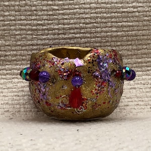 May include: A small, gold-colored ceramic pot with red, purple, and iridescent glitter. The pot has a beaded trim with red, purple, and iridescent beads.