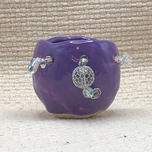 May include: A small, round, purple ceramic vase with a glossy finish. The vase is adorned with three decorative beaded accents, each featuring clear, iridescent beads. The vase has a small opening at the top and sits on a white surface.