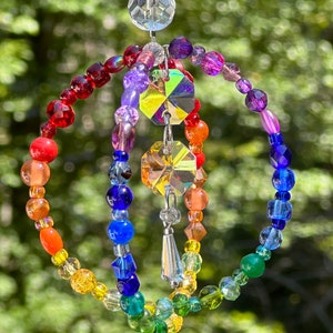 May include: A colorful suncatcher with a rainbow of beads and crystal accents. The design features three concentric beaded rings in shades of red, orange, yellow, green, blue, and purple, with a central crystal prism and dangling crystal pendant.