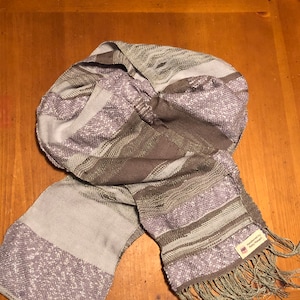 May include: A gray and white woven scarf with a fringe. The scarf has a label that reads "Handwoven by Patty Magee".