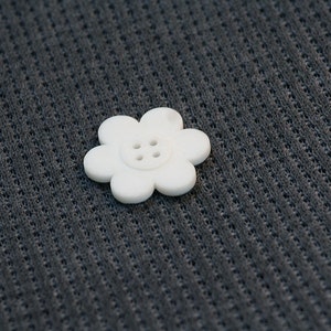 May include: A white flower-shaped button with four holes in the centre. The button is on a dark grey textured fabric.