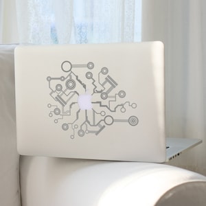 Op de afbeelding: Een zilveren laptop met een grijze sticker met een print van een circuitbord op de achterkant. De sticker heeft een gestileerd ontwerp van verbonden cirkels en lijnen.