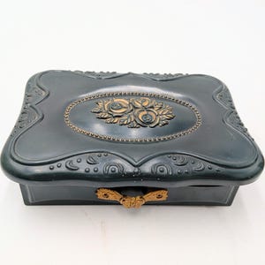 May include: A dark teal jewelry box with an ornate design. The box features a gold-colored floral embellishment on the lid and a matching handle. The box has a rectangular shape with rounded edges and decorative detailing.