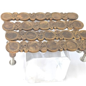 May include: Antique brass-toned drawer pulls with a circular design. Each pull features a series of connected circles and small decorative elements. The pulls are mounted on silver-colored screws, and are displayed on a clear, geometric stand.