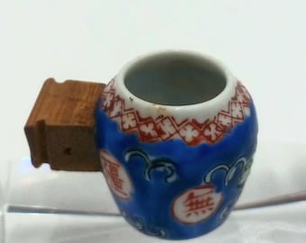 May include: A small, blue ceramic jar with white and red floral patterns and Chinese characters. The jar has a wooden stopper attached to the side. The jar is on a white surface.