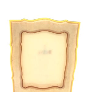 May include: A vintage-style picture frame with a yellow border and a light brown interior. The frame has a decorative, scalloped edge and a rectangular opening for a photo or artwork. The frame is made of a light-colored material.