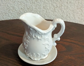 May include: A white ceramic creamer with a decorative floral design and a matching saucer. The creamer has a curved handle and a spout for easy pouring. The set is made of ceramic and is suitable for serving cream or milk.