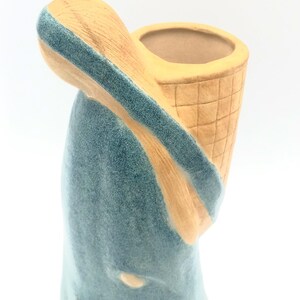 May include: A ceramic figurine of a woman carrying a basket on her back. The figurine is glazed in a light blue color with a tan base.