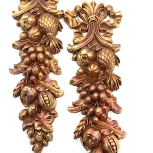 May include: A pair of gold-toned wall decor pieces featuring a stylized design of fruit and leaves. The pieces are made of a textured material and have a vintage look.