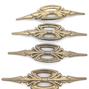 May include: Four identical elongated metal drawer pulls in a muted brass finish. Each pull features a central oval frame with a horizontal bar, flanked by pointed, spear-like ends. The design includes a small ribbed detail at the center.