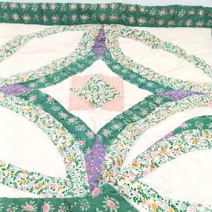 May include: A white and green patchwork quilt with a floral pattern. The quilt is made up of squares and circles, and the fabric is a mix of white, green, pink, and purple.