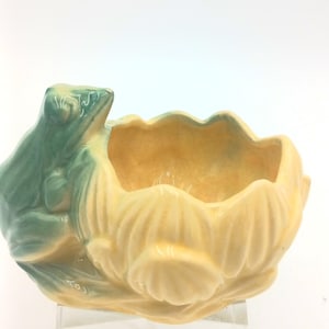 May include: A ceramic frog planter in shades of green and yellow. The frog is perched on the side of a flower-shaped container. The planter is designed with a glossy finish and is suitable for small plants or decorative items.