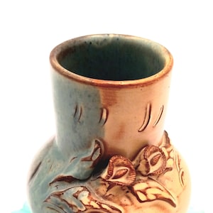 May include: A handcrafted ceramic vase with a two-tone design. The upper portion is a light brown, transitioning to a teal blue base. The vase features carved floral and leaf details, adding a touch of nature-inspired artistry to the piece.