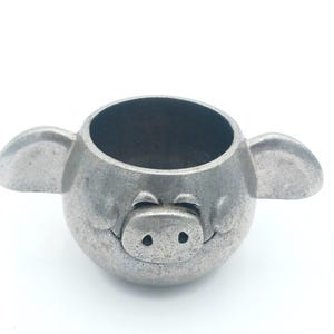 May include: A small, silver pig-shaped cup with a round body and two small ears. The pig has a smiling face with two small nostrils and a closed mouth.