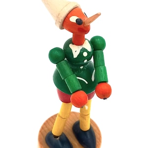 May include: A wooden puppet with a green shirt, red pants, and a white hat. The puppet has a long nose and is standing on a wooden base.