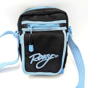 May include: A black crossbody bag with light blue trim and an adjustable light blue shoulder strap. The front features a zippered pocket and the embroidered white text Roxy with a diamond logo. The main compartment also has a zipper closure.