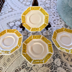 May include: A collection of vintage white and yellow octagonal saucers. Each saucer features a yellow floral pattern around the rim and a central indentation for a teacup. The saucers are arranged on a white lace tablecloth.