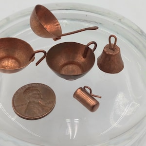 May include: Miniature copper kitchenware set including a ladle, two pans, a bell, and a small pitcher. A penny is included for size comparison. The items are made of copper and have a shiny, metallic finish.