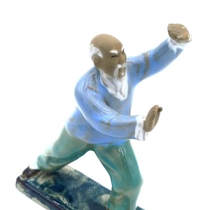 May include: A ceramic figurine of an elderly man in a martial arts pose. The figure wears a light blue top and teal pants. The figurine is on a rectangular base with a dark blue glaze.