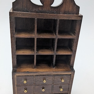 May include: Miniature wooden cabinet with a dark brown finish. The cabinet features a top section with multiple small cubby shelves and a lower section with three rows of drawers. Each drawer has a small, round, gold-colored knob.