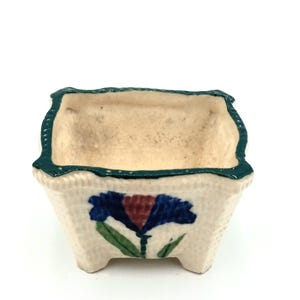 May include: A small, square ceramic planter with a textured, off-white exterior. The rim is trimmed with a dark green border. A blue and red floral design is painted on the front. The planter has a beige interior and is elevated by small feet.
