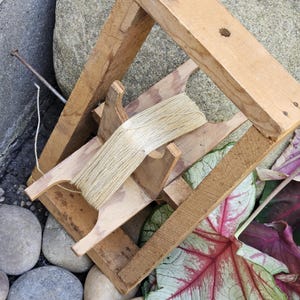 May include: A wooden yarn swift with a skein of beige yarn. The swift is made of light-colored wood and has a rectangular frame. The yarn is wound around the swift's arms. The swift is resting on a bed of rocks and leaves.