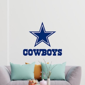 May include: A navy blue star and the word "COWBOYS" are displayed on a white wall. Below, a gray sofa is adorned with teal and orange pillows. A small table holds decorative items.