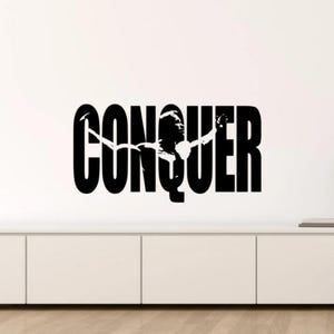 May include: Black and white wall decal with the word "CONQUER" in bold letters. The word is stylized with a silhouette of a person in a flying pose.