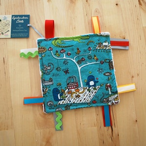 Könnte beinhalten: Eine türkisblaue Babysinnenstirnband mit einem Baum, Tieren und einer Picknickszene. Die Decke hat mehrere bunte Bänder an den Rändern.