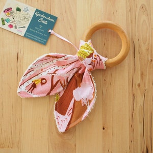 May include: A pink and brown fabric teether with a wooden ring attached. The fabric has a woodland pattern with a girl in a yellow dress walking through the trees. The teether is tied in a bow shape. The tag attached to the teether reads "Epideron Little" and includes the words "Baby Clothes, Teething & Sensory Toys".